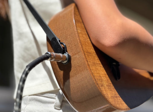 Close-up of a person holding an acoustic electric ukulele with a strap lock and amp cable.